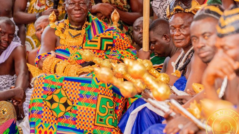 A chief seated among elders, wearing kente and gold regalia.